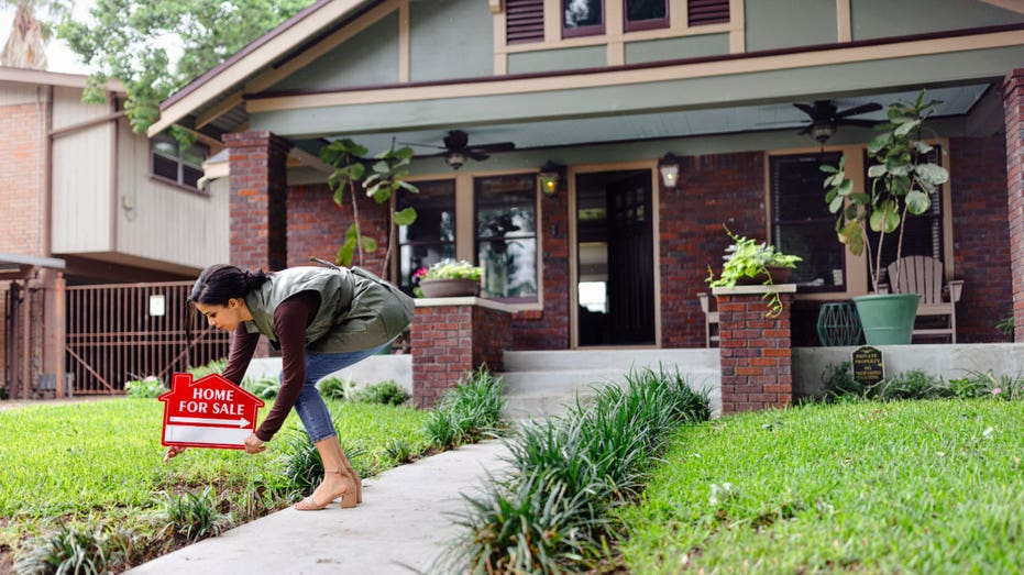 A real estate agent holds a "for sale" sign in front of a house.